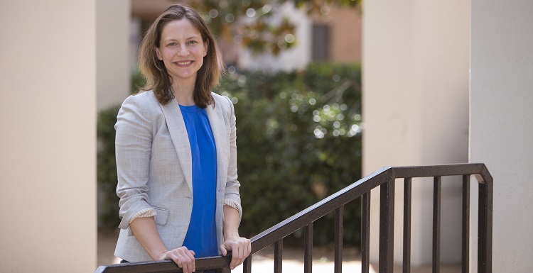 Claire Cage wears a light grey blazer and blue shirt, and stands with her hands on a staircase railing outside the Humanities Building.